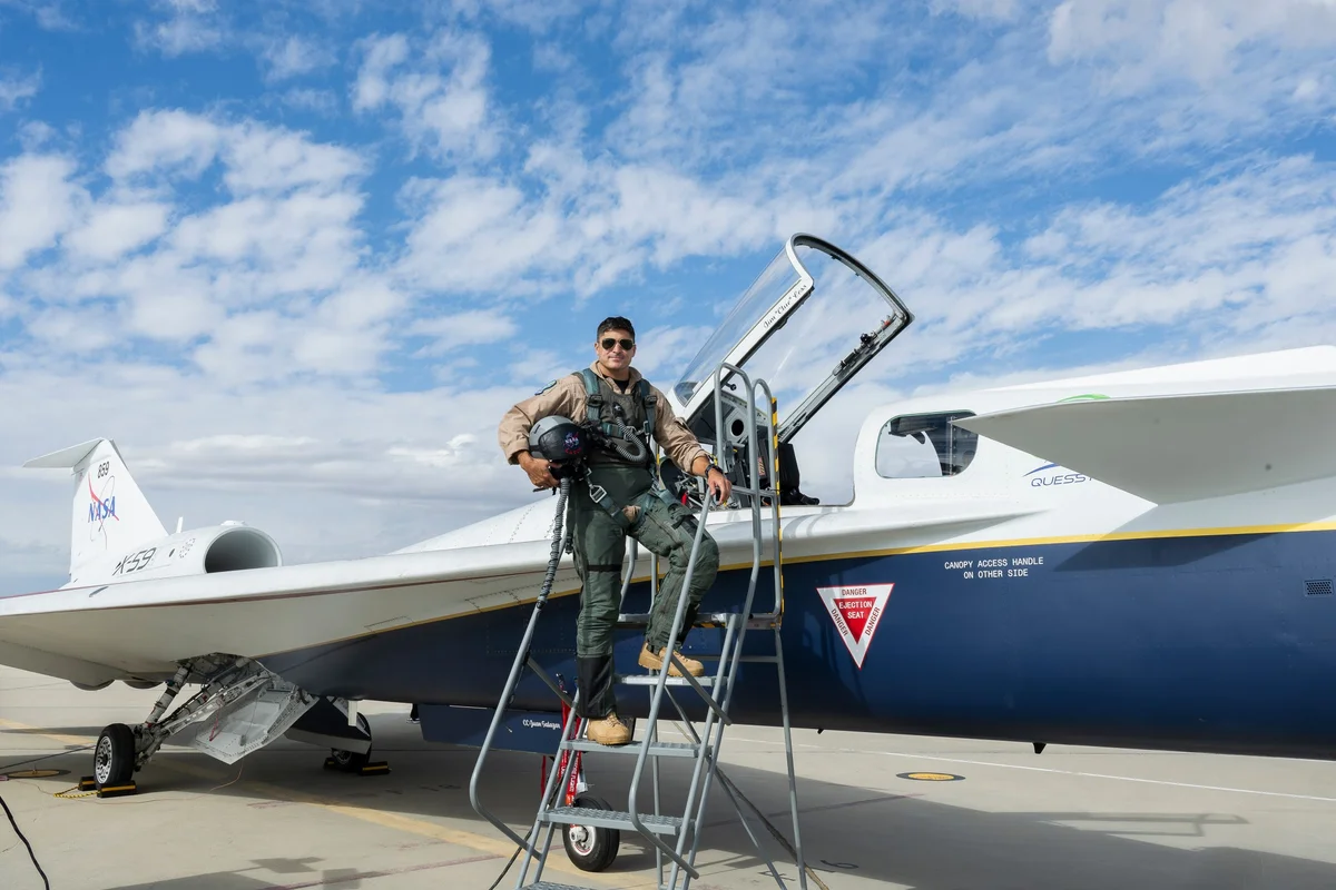 NASA test pilot Mike Lippert stands on a gray metal ladder before getting into the X-59 plane. He is holding his helmet under his right arm. The cockpit of the plane is open.